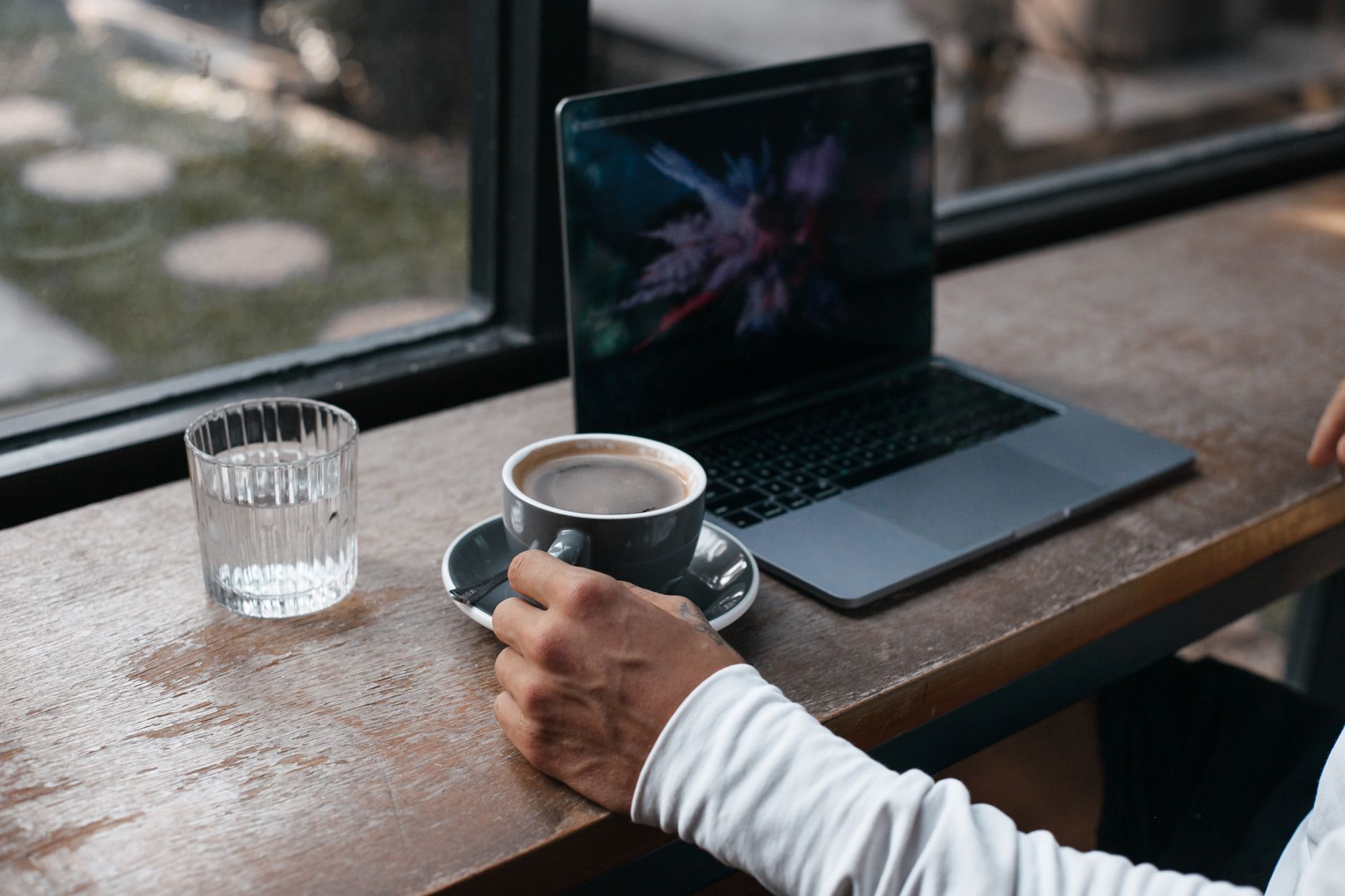 Hand holding a coffee cup next to a laptop on a wooden table by a window, a peaceful home work setting.