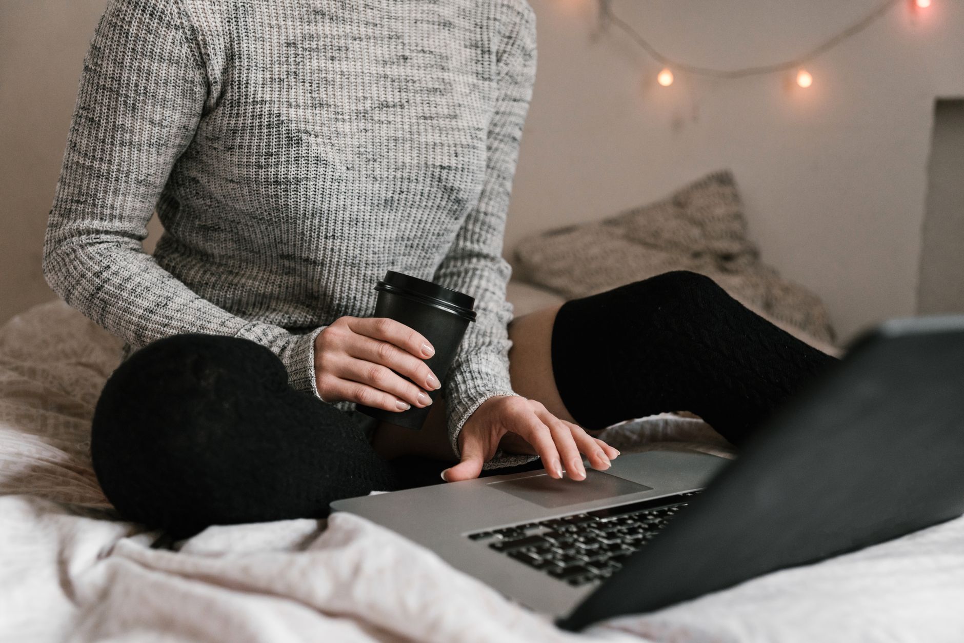 A woman in a sweater working with coffee and a laptop on a bed, a relaxed home work setting.