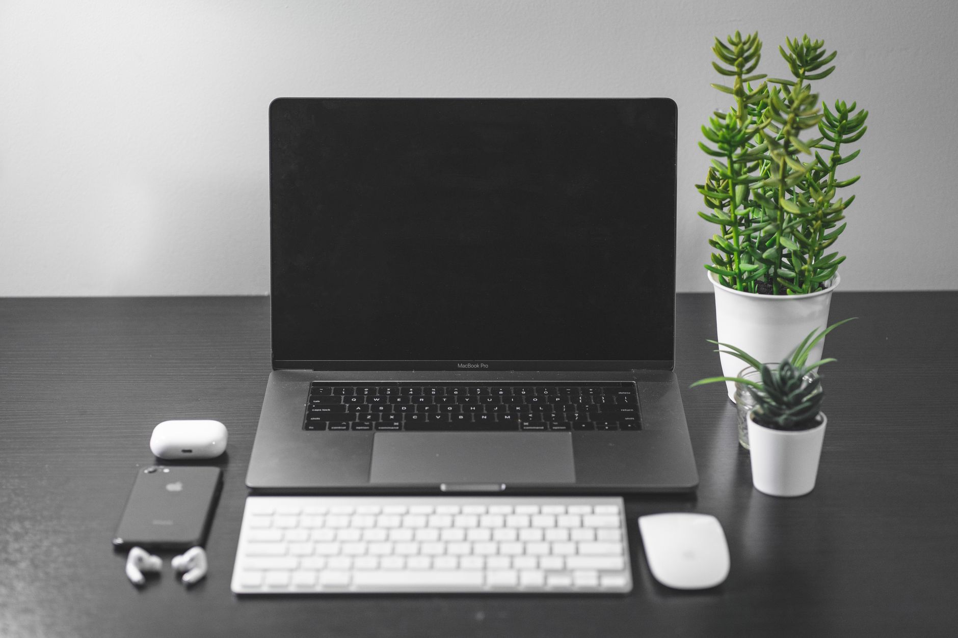 Secretária escura com um portátil aberto, teclado e rato brancos, telemóvel, auriculares e duas plantas em vasos brancos